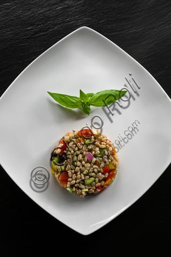 Photograph of a serving of spelt grain salad with cherry tomatoes, fresh basil leaves, and black olives in a white bowl.