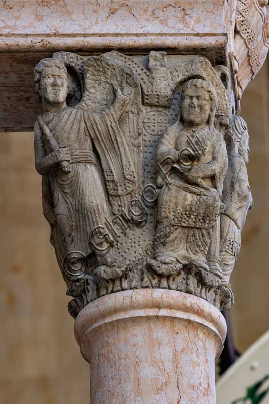 Fidenza, Duomo (Cathedral of San Donnino), facade, main portal, prothyrum: the left capital with “Stories of the Virgin Mary”.  the “Annunciation”.the Archangel Gabriel, three-quarters to the left, has an object in his right hand and blesses with his left; above is the inscription GABRIHEL (Gabriel = Gabriel). The Madonna is seated, dressed in a surplice over her tunic and with a veil on her head; in her right hand she has a sphere (perhaps a ball of thread) and in her left hand she holds two spindles in her lap; above is the inscription SCAMARIA (Sancta Maria = Holy Mary).Work by Benedetto Antelami and workshop.