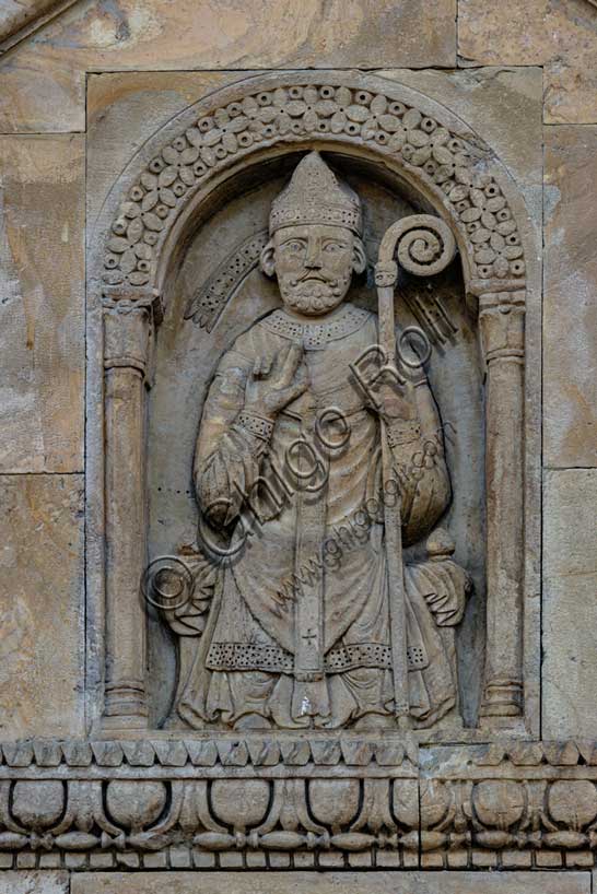 Fidenza, Duomo (Cattedrale di San Donnino), facciata, timpano del portale destro: “figura di Vescovo”. il personaggio è seduto su un faldistorio, vestito di camice, cotta e casula, con mitria e pastorale. Tradizionalmente nella figura è stato riconosciuto l’Arciprete della chiesa di S. Donnino, ma il pallio riservato sin dal medioevo agli arcivescovi metropoliti e ai primati, fa dubitare di questa interpretazione. Opera di Benedetto Antelami e bottega.