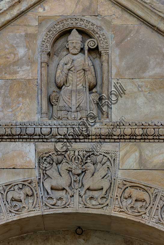 Fidenza, Duomo (Cattedrale di San Donnino), facciata, timpano del portale destro: “figura di Vescovo”. il personaggio è seduto su un faldistorio, vestito di camice, cotta e casula, con mitria e pastorale. Tradizionalmente nella figura è stato riconosciuto l’Arciprete della chiesa di S. Donnino, ma il pallio riservato sin dal medioevo agli arcivescovi metropoliti e ai primati, fa dubitare di questa interpretazione. Opera di Benedetto Antelami e bottega.