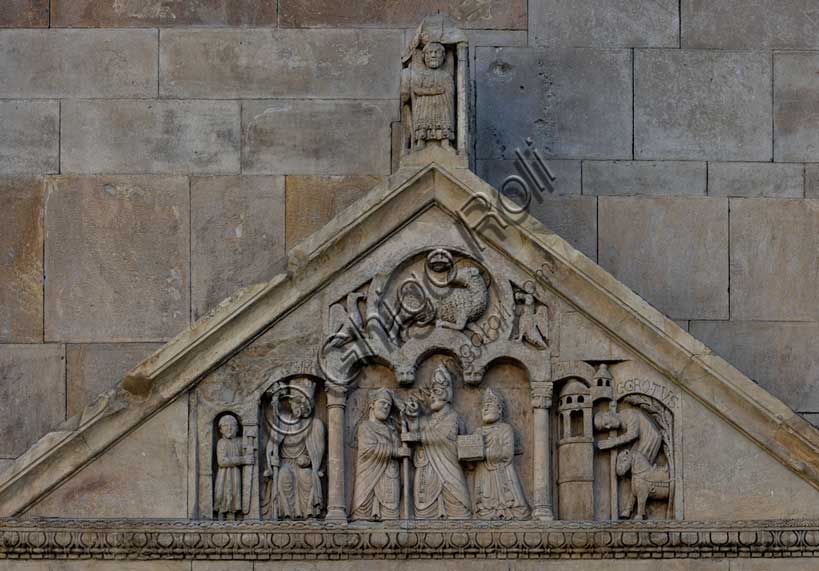 Fidenza, duomo (Cattedrale di San Donnino), portale sinistro, timpano. Nel frontone si leggono, da sinistra, tre scene: L'imperatore Carlo Magno, con un armigero alla sua destra;Papa Adriano II che porge all'arciprete di San Donnino la mitria e il pastorale (simboli della dignità episcopale); il miracolo dell'ammalato guarito (miracolo compiuto da San Donnino).Nell’acroterio sul timpano, é raffigurato un personaggio, probabilmente Oberto II Pallavicino condottiero posto da Federico II di Svevia alla guida della città dal 1249 al 1268.Opera di Benedetto Antelami e bottega.