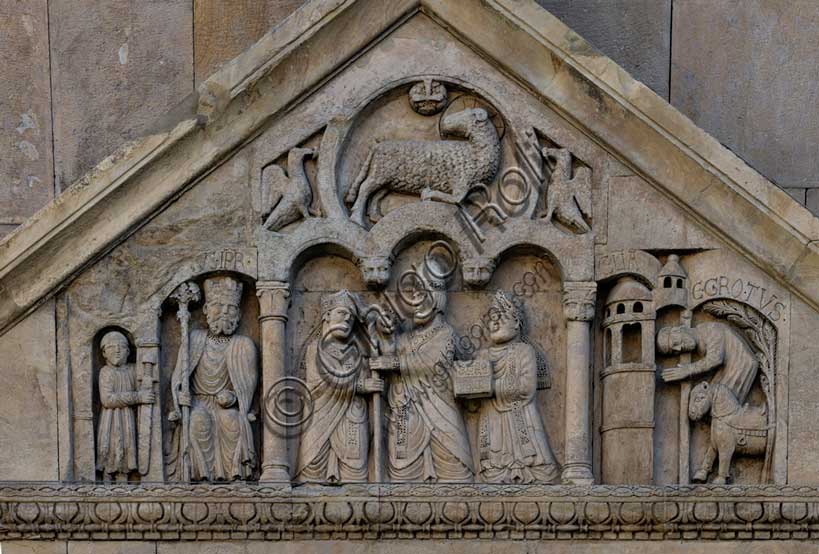 Fidenza, duomo (Cattedrale di San Donnino), portale sinistro, timpano. Nel frontone si leggono, da sinistra, tre scene: L'imperatore Carlo Magno, con un armigero alla sua destra;Papa Adriano II che porge all'arciprete di San Donnino la mitria e il pastorale (simboli della dignità episcopale); il miracolo dell'ammalato guarito (miracolo compiuto da San Donnino).Nell’acroterio sul timpano, é raffigurato un personaggio, probabilmente Oberto II Pallavicino condottiero posto da Federico II di Svevia alla guida della città dal 1249 al 1268.Opera di Benedetto Antelami e bottega.