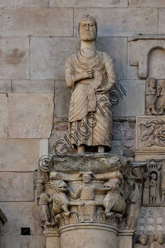 Fidenza, duomo (Cattedrale di San Donnino), sculture tra il portale maggiore e quello di sinistra. Particolare di “San Simone Apostolo che mostra la via di Roma ai pellegrini”; la statua tiene fra le mani un filatterio che si svolge verso il basso sul quale è incisa a lettere capitali l’iscrizione: S.IMO / APLS / EUDI / ROMA / SCDS / MON / A[…]RAT / HAC / VIAM (Simon Apostolus eundi Romam demonstrat hanc viam).La statua sormonta il semicapitello con “Daniele nella fossa dei leoni”.Opera di Benedetto Antelami e bottega.