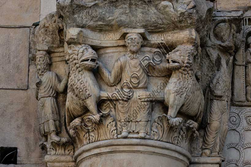 Fidenza, duomo (Cattedrale di San Donnino), sculture tra il portale maggiore e quello di sinistra. Particolare del semicapitello con “Daniele nella fossa dei leoni”. Il semicapitello figurato che raffigura nella parte frontale la scena biblica – di simbologia cristologica – del profeta Daniele che gettato nella fossa è attorniato dai leoni ammansiti. Sua lato destro del semicapitello è raffigurato il profeta Abacuc con il cibo per Daniele e sul lato sinistro l’angelo che lo portò (Dn 14,33-38). Sull’abaco del semicapitello è incisa a lettere capitali l’iscrizione: […]C DANIEL IVSTUS INLA EST CV LEONV[M] ([Abacu]c . Daniel iustus in lacu est cum leonum).Opera di Benedetto Antelami e bottega.