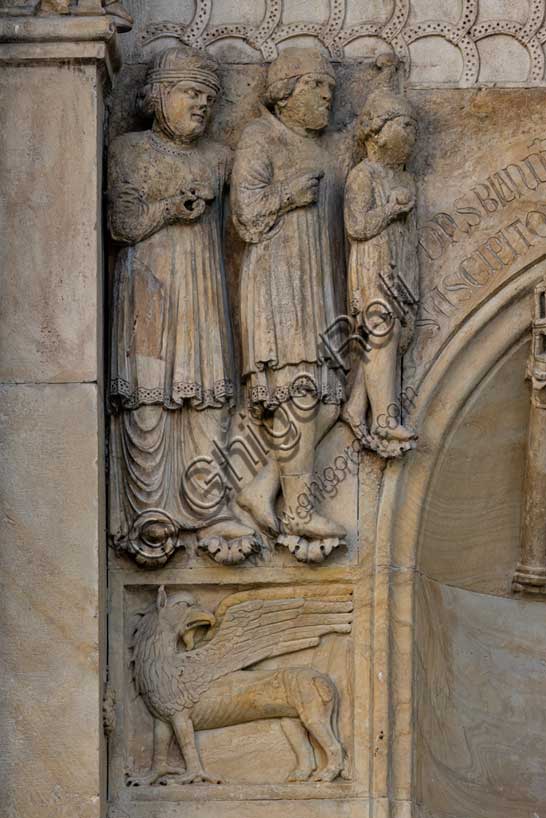 Fidenza, duomo (Cattedrale di San Donnino), sculture tra il portale maggiore e quello di sinistra vicino alla nicchia con il “Re David” di Benedetto Antelami.Particolare con le figura di un grifo sormontata dal bassorilievo raffigurante verosimilmente una famiglia di pellegrini ricchi in viaggio per Roma. 