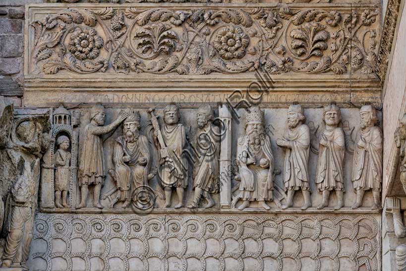 Fidenza, duomo (Cattedrale di San Donnino), sculture tra il portale maggiore e quello di sinistra vicino alla nicchia con il “Re David” di Benedetto Antelami.“Storie di San Donnino” con il primo e secondo episodio: Nel pannello a sinistra, la scena è rappresentata all’esterno d’un edificio sotto il quale si trova una guardia armata. Donnino, cubiculario di Massimiano Erculeo, stando alla destra dell’imperatore, gli pone la corona sul capo; alla sinistra di Massimiano il Portaspada dimostra che la scena costituisce un momento dell’esercizio del potere sovrano di giurisdizione, probabilmente nei confronti del personaggio posto all’estrema destra. Nella cornice attigua posta sopra è incisa a lettere capitali l’iscrizione: IR . ABEATO . DOMNINO CORONATVR (Imperator a Beato Domnino coronatur = L’Imperatore è incoronato dal beato Donnino).Nel pannello a destra la distinzione dalla scena precedente è resa evidente, oltre che dal diverso colore della pietra, da una torre merlata poligonale oltre la quale è seduto l’imperatore in maestà con i segni regali, a fianco del quale Donnino sta in piedi di profilo con le mani giunte in atto di “istanza” e dietro di lui due altri personaggi – vestiti come lui – alzano la mano in segno di assenso. Sulla cornice contigua superiore è incisa a lettere capitali l’iscrizione: LICENCIA . ACEPTA . DEO . SERVIREDECRE[VIT] (Licencia acepta Deo servire decrevit = Ricevuto il permesso, decise di servire Dio).