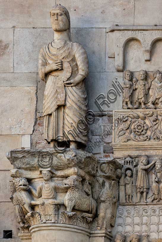 Fidenza, duomo (Cattedrale di San Donnino), sculture tra il portale maggiore e quello di sinistra. Particolare di “San Simone Apostolo che mostra la via di Roma ai pellegrini”; la statua tiene fra le mani un filatterio che si svolge verso il basso sul quale è incisa a lettere capitali l’iscrizione: S.IMO / APLS / EUDI / ROMA / SCDS / MON / A[…]RAT / HAC / VIAM (Simon Apostolus eundi Romam demonstrat hanc viam).La statua sormonta il semicapitello con “Daniele nella fossa dei leoni”.Opera di Benedetto Antelami e bottega.