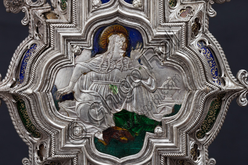 Florence, Museum of the Opera del Duomo (Museum of Florence Cathedral of Santa Maria del Fiore): “The Cross of the Treasure of St. John: the recto.”Detail of the upper part with the figure of a saint or Madonna.The Cross is a splendid example of Renaissance jewelery, sculpted in silver foil, chiseled and enameled by Betto di Francesco Betti, Antonio del Pollaiolo and Miliano di Domenico Dei between 1457 and 1459 approximately. It was commissioned to complete the silver altar. The Cross-monstrance is made up of three distinct main parts: the Crucifix, the stem and the base. 