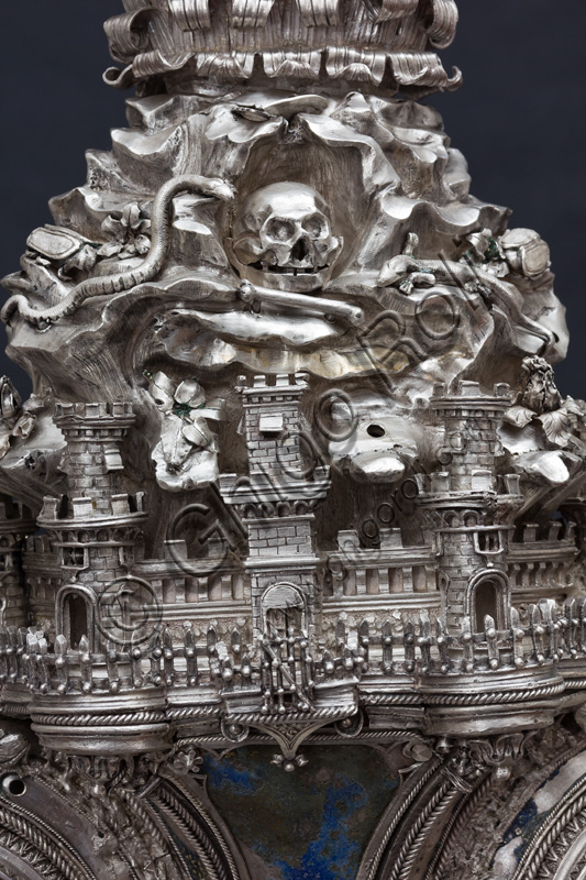 Florence, Museum of the Opera del Duomo (Museum of Florence Cathedral of Santa Maria del Fiore): “The Cross of the Treasure of St. John: the recto.”Detail of the central part with buildings of a city, a snake and a skull.The Cross is a splendid example of Renaissance jewelery, sculpted in silver foil, chiseled and enameled by Betto di Francesco Betti, Antonio del Pollaiolo and Miliano di Domenico Dei between 1457 and 1459 approximately. It was commissioned to complete the silver altar. The Cross-monstrance is made up of three distinct main parts: the Crucifix, the stem and the base. 