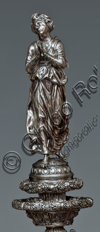 Firenze, Museo dell'Opera del Duomo (Santa Maria del Fiore): “La croce del tesoro di S. Giovanni: il verso”. Particolare di “statua di angelo orante”.La Croce è uno splendido esempio di oreficeria rinascimentale, scolpita in lamina d’argento, cesellata e smaltata da Betto di Francesco Betti, Antonio del Pollaiolo e Miliano di Domenico Dei tra il 1457 e il 1459 circa. Venne commissionata a completamento dell’altare d’argento. La Croce-ostensorio è composta da tre parti principali distinte: il Crocifisso, il fusto e la base. 