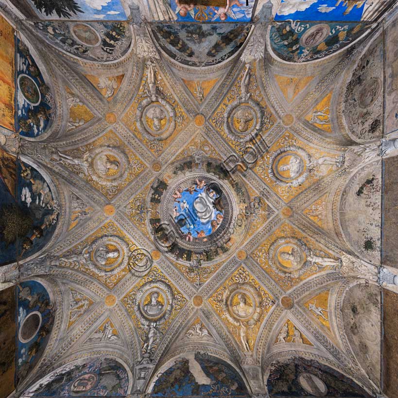 Ducal Palace: vault of the “Bridal Chamber or Camera Picta", which is a room located in the north-east tower of the Castle of San Giorgio in Mantua. It is famous for the cycle of frescoes that covers its walls, a masterpiece by Andrea Mantegna, created between 1465 and 1474.Detail of the vault illusionistically divided into painted rib vault and pendentives. Fake ribs divide the space into regular figures, with a golden background and monochrome paintings.In the center is the famous oculus. It is a round open illusionistically towards the sky, which was supposed to recall the famous oculus of the Pantheon. In the oculus you can see a balustrade from which a court lady leans out, accompanied by her black servant, a group of maids, a dozen putti, a peacock and a vase, against the background of a blue sky. To reinforce the impression of the open oculus, Mantegna painted some cherubs dangerously poised clinging to the inside of the frame, with dizzying glimpses of their plump little bodies.The oculus is enclosed by a circular garland, which is in turn enclosed in a square of fake ribs. At the meeting points there are golden medallions. Arranged around the square are eight lozenges with a golden background, each containing a circular garland enclosing a portrait of one of the first eight Roman emperors.