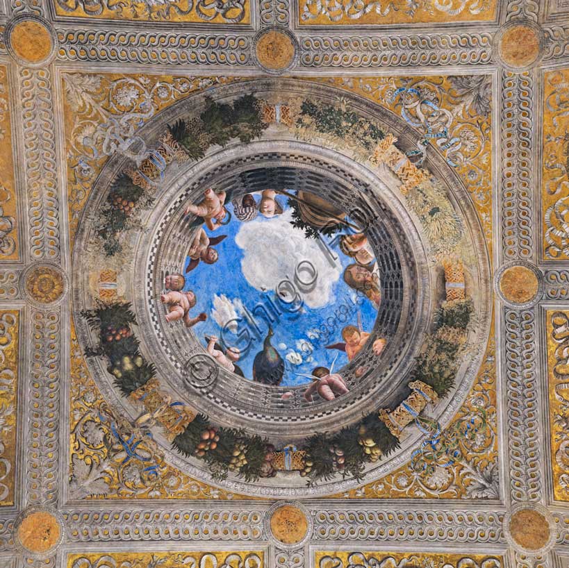 Ducal Palace: vault of the “Bridal Chamber or Camera Picta", which is a room located in the north-east tower of the Castle of San Giorgio in Mantua. It is famous for the cycle of frescoes that covers its walls, a masterpiece by Andrea Mantegna, created between 1465 and 1474.Detail of the vault illusionistically divided into painted rib vault and pendentives. The space is divided into regular figures, with a golden background and monochrome paintings.In the center is the famous oculus. It is a round open illusionistically towards the sky, which was supposed to recall the famous oculus of the Pantheon. In the oculus you can see a balustrade from which a court lady leans out, accompanied by her black servant, a group of maids, a dozen putti, a peacock and a vase, against the background of a blue sky. To reinforce the impression of the open oculus, Mantegna painted some cherubs dangerously poised clinging to the inside of the frame, with dizzying glimpses of their plump little bodies.